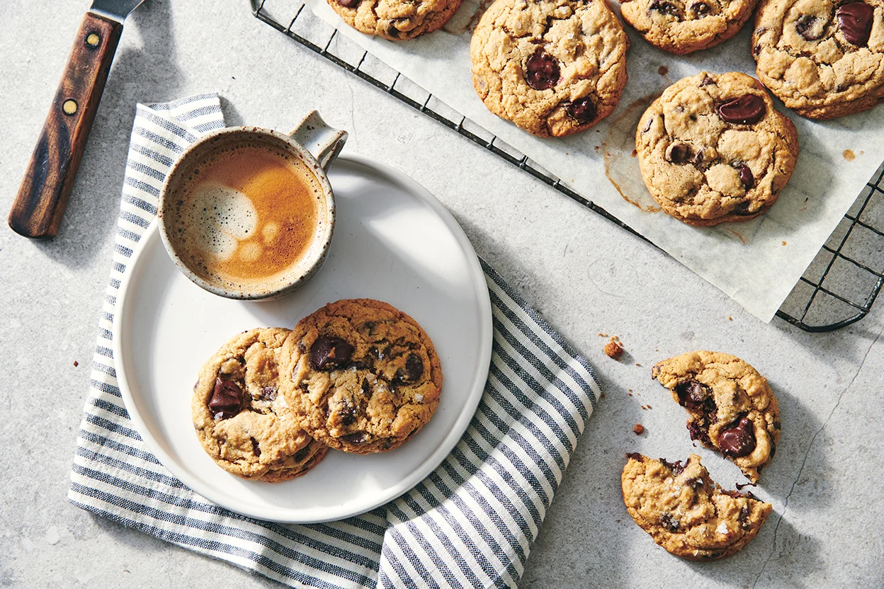 A batch of soft chocolate chip cookies cooling on a wire rack with a baking sheet nearby on a kitchen counter