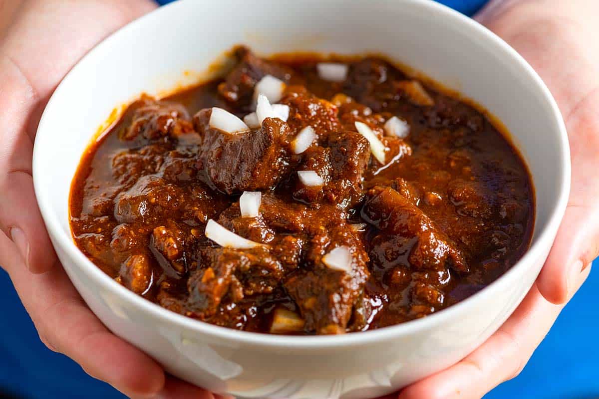 A bowl of Texas-style chili with a light creamy swirl on top, garnished with sliced jalapeño and chopped cilantro on a wooden table