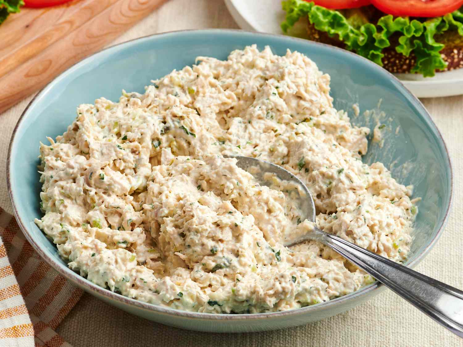 A bowl of creamy chicken salad with celery and herbs on a wooden counter, with a toasted sandwich on a plate beside it
