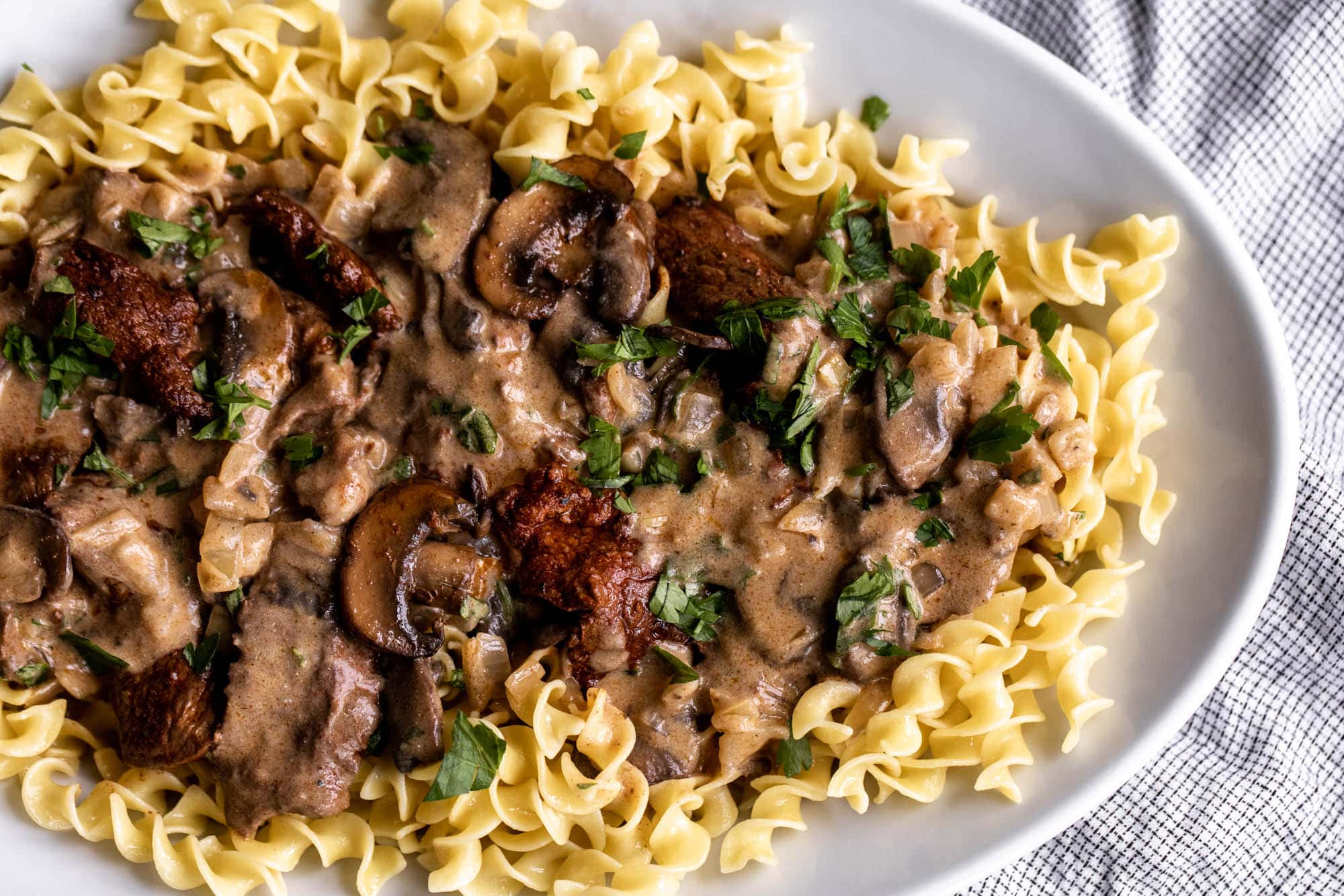 A bowl of creamy steak stroganoff spooned over egg noodles with mushrooms and a sprinkle of chopped parsley on top
