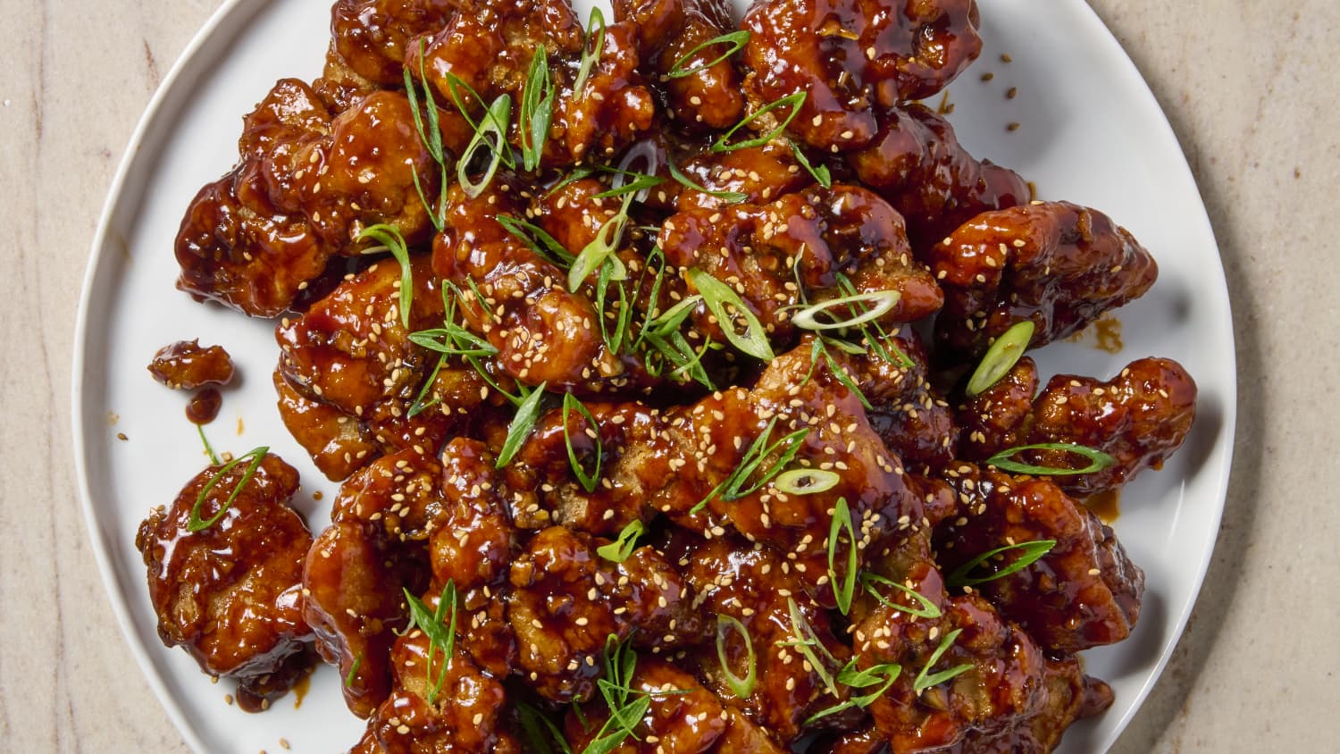 A bowl of crispy Asian chicken pieces coated in a glossy caramel-brown sauce, topped with sliced scallions and sesame seeds, served over steamed white rice on a wooden table