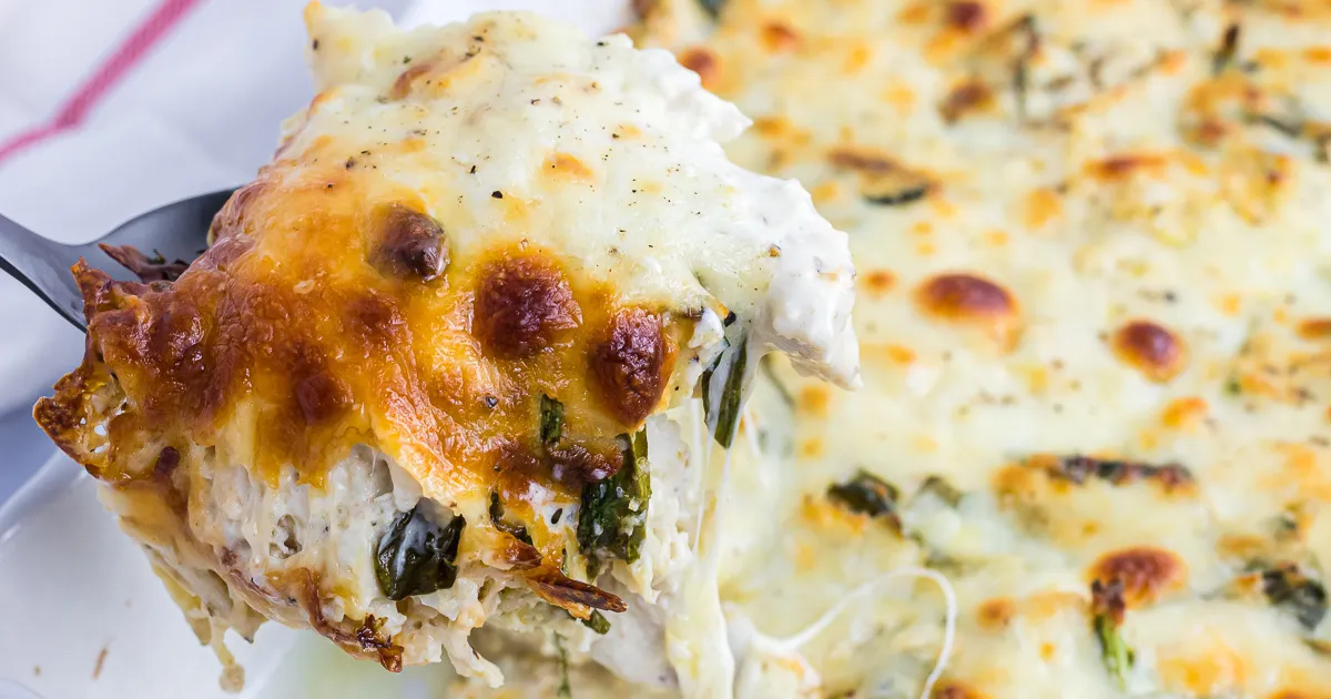 A bubbling keto chicken Alfredo bake in a white casserole dish with a golden cheese top, fresh parsley sprinkled over, sitting on a kitchen counter with a serving spoon nearby