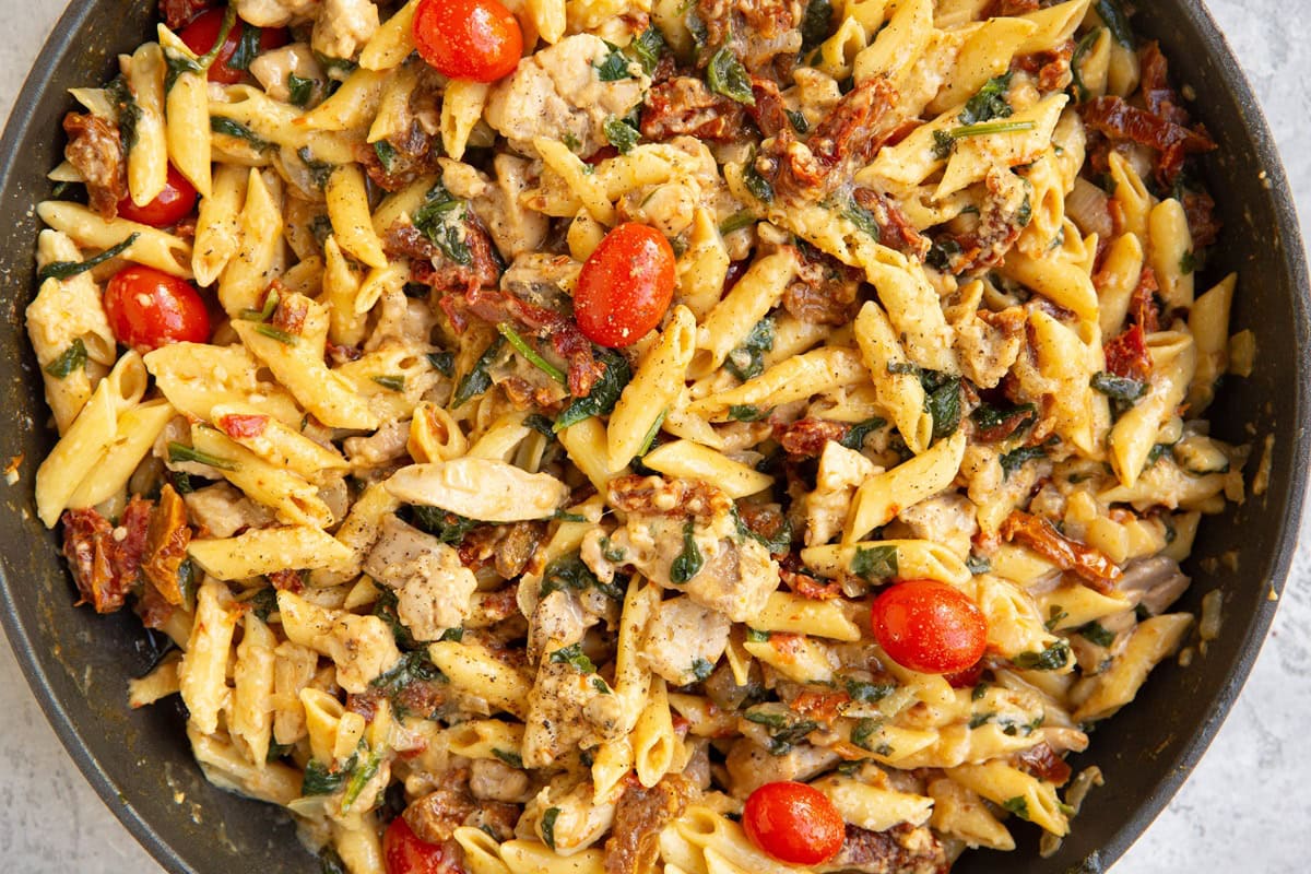 A cast iron skillet filled with rustic chicken and pasta, with seared chicken thighs nestled into saucy pasta and cherry tomatoes, garnished with parsley on a wooden table