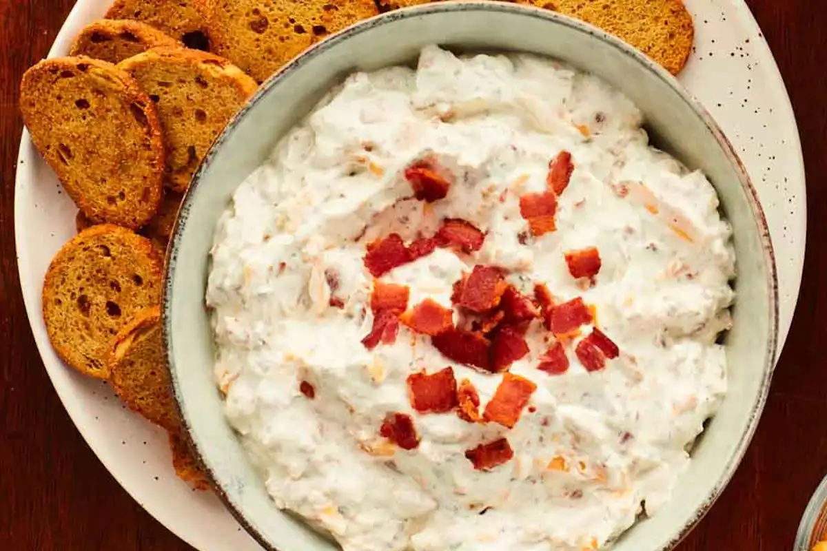 A ceramic bowl of creamy ranch dip topped with chopped chives on a wooden table, surrounded by carrot sticks, celery, and ridged potato chips