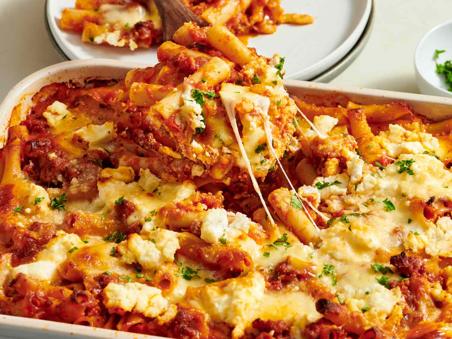 A deep baking dish filled with baked ziti topped with melted mozzarella and browned cheese spots, sitting on a wooden table with a serving spoon