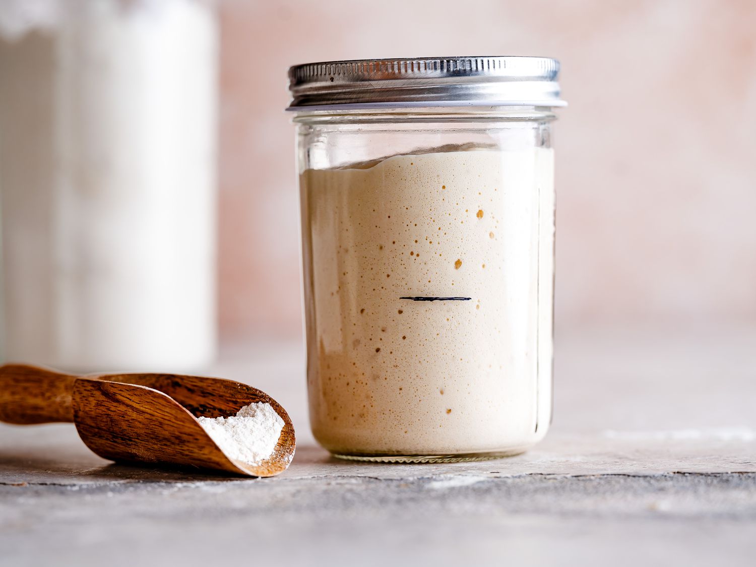 A glass jar of sourdough starter with visible bubbles sitting on a kitchen counter next to a spoon and a bag of flour