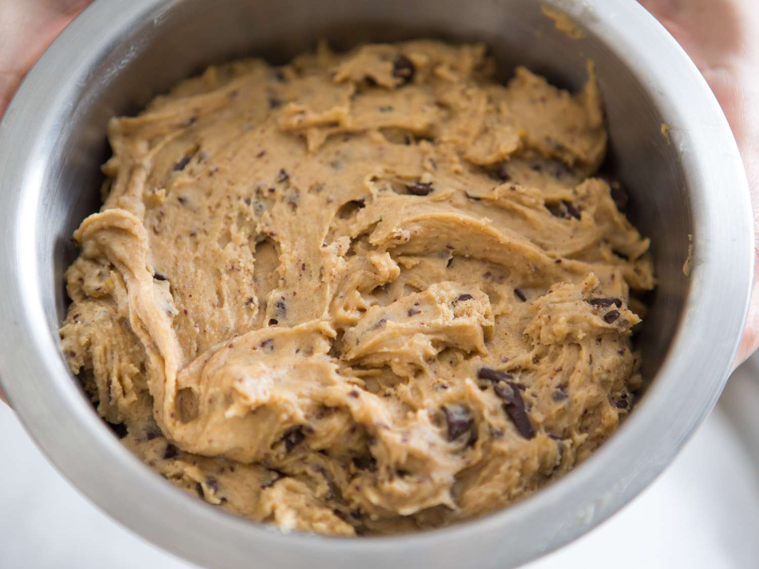 A glass mixing bowl filled with chocolate chip cookie dough, with a wooden spoon resting in the dough on a bright kitchen counter