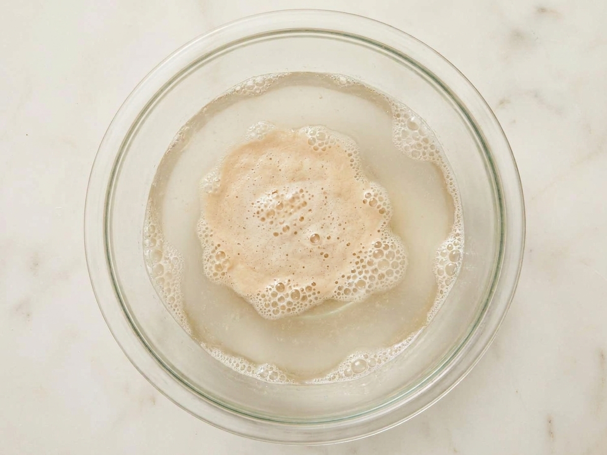 A glass mixing bowl with warm water and yeast blooming on the surface, small bubbles forming around the edges