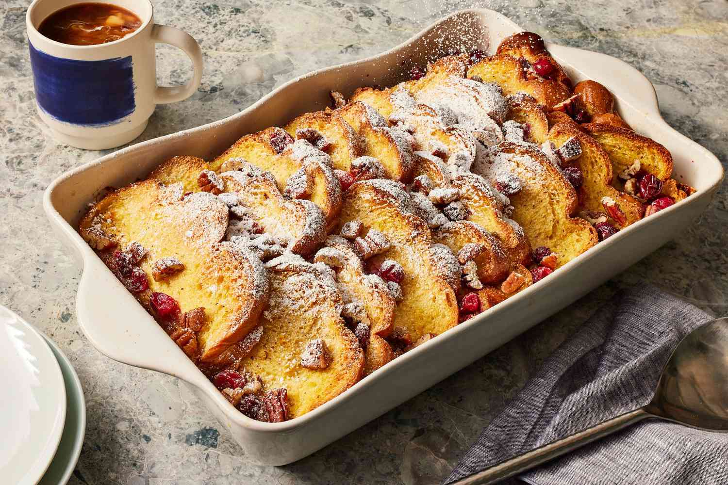A golden baked French toast casserole in a ceramic baking dish on a wooden table with a serving spoon nearby