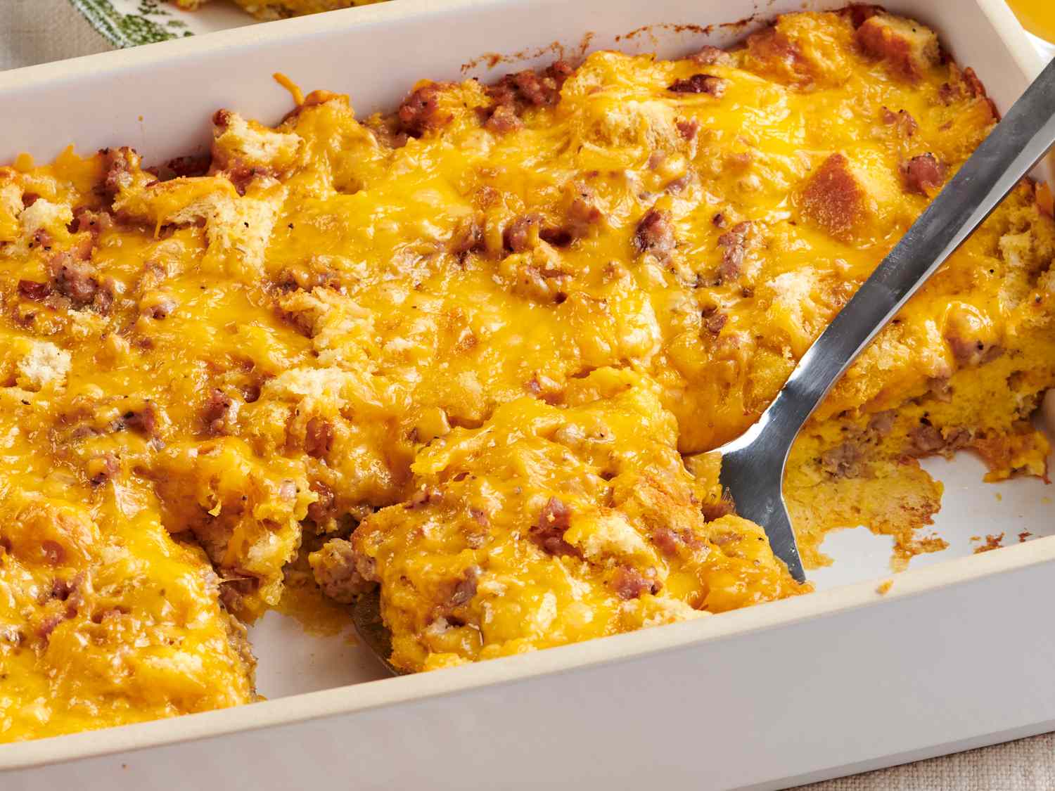 A golden-brown breakfast casserole in a white ceramic baking dish on a kitchen counter, with a serving spoon and steam rising