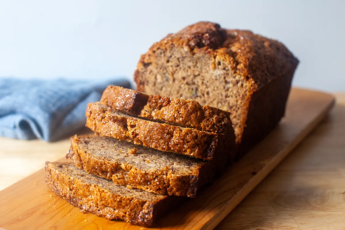 A golden brown loaf of banana bread cooling on a wire rack on a kitchen counter, with a few banana slices visible on top