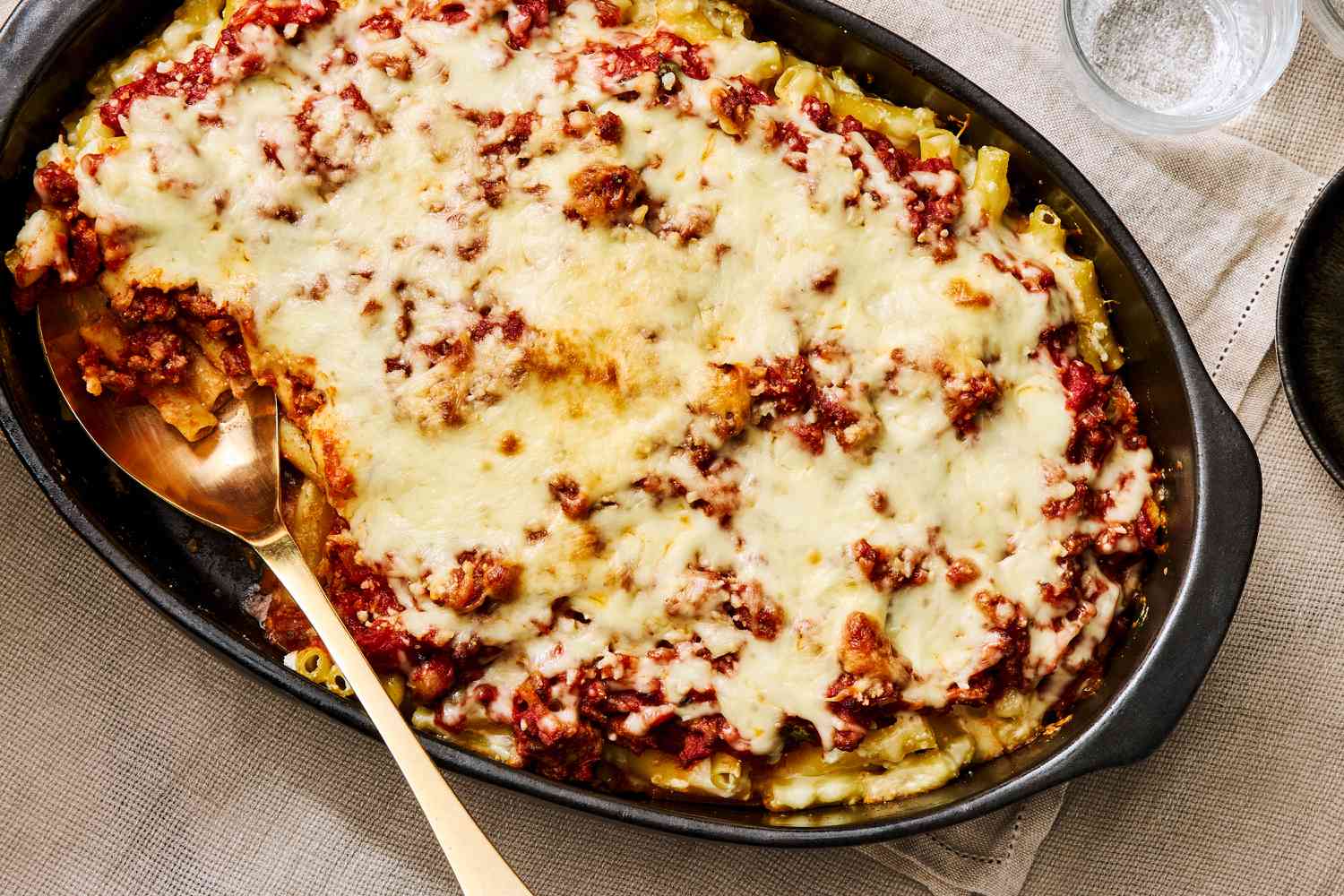 A golden, bubbly pan of baked ziti with browned mozzarella and a sprinkle of parsley, photographed on a kitchen counter with a serving spoon