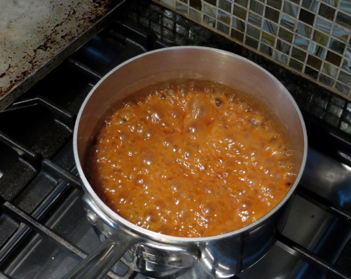 A heavy-bottomed saucepan on a stovetop with pecans being coated in bubbling caramel as a wooden spoon stirs