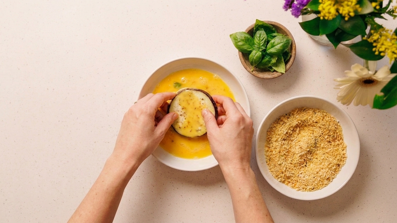 A home cook's hands dipping an eggplant slice into beaten egg beside a bowl of seasoned breadcrumbs on a kitchen counter