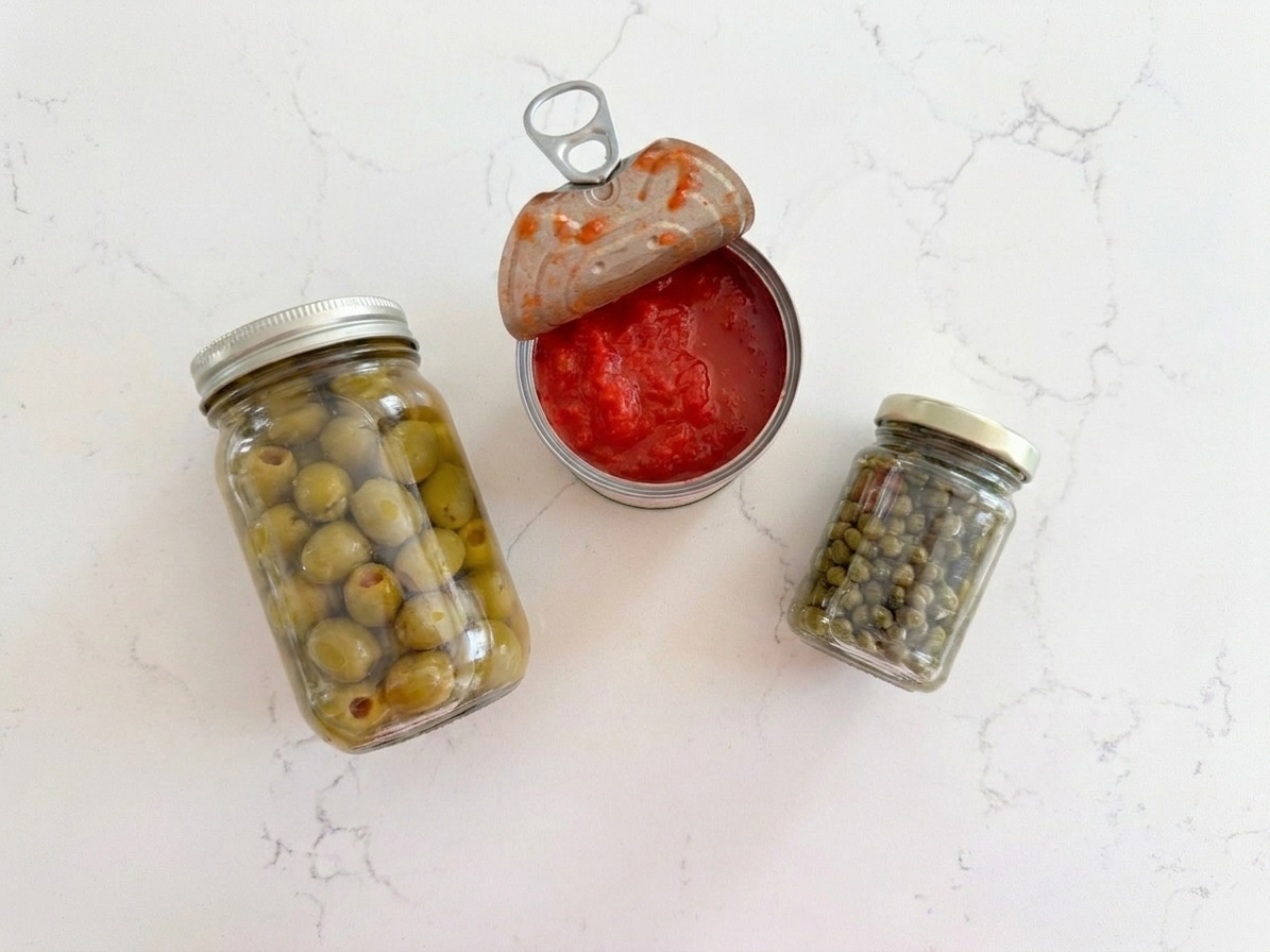 A jar of olives, a jar of capers, and an open can of tomatoes on a countertop