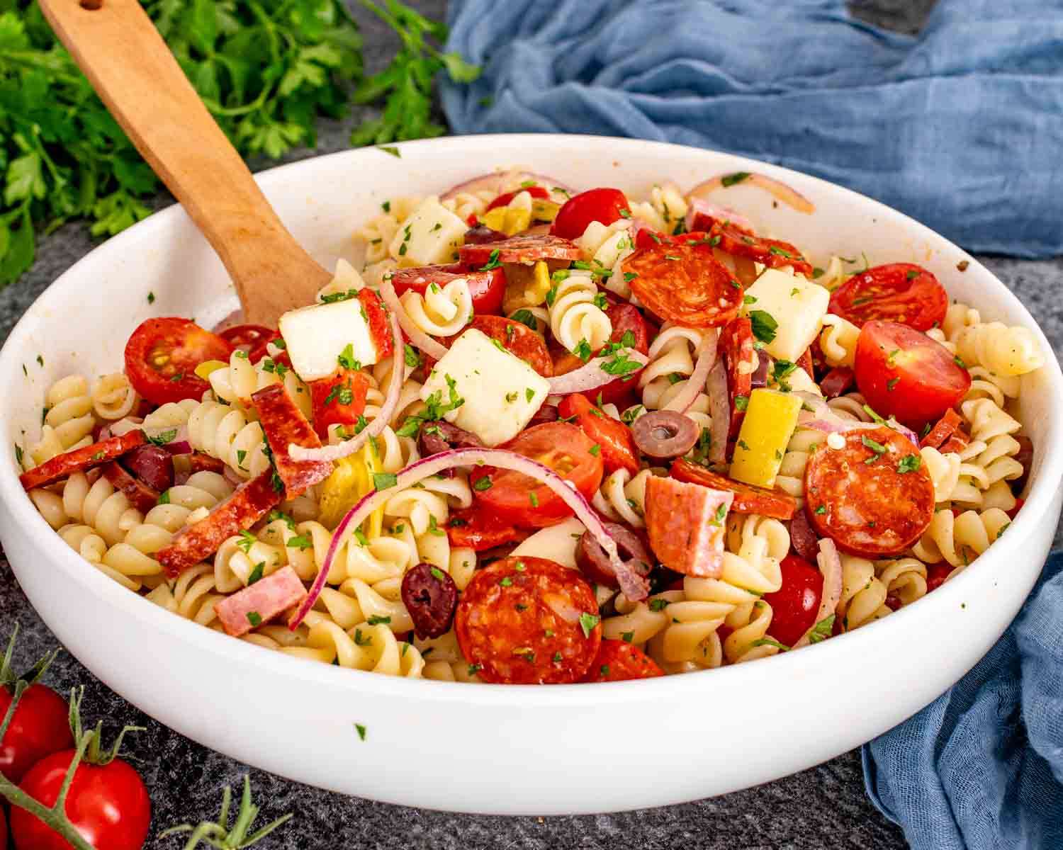 A large ceramic bowl filled with fresh pasta salad with cherry tomatoes, cucumber, basil, mozzarella, and a glossy vinaigrette on a wooden kitchen counter