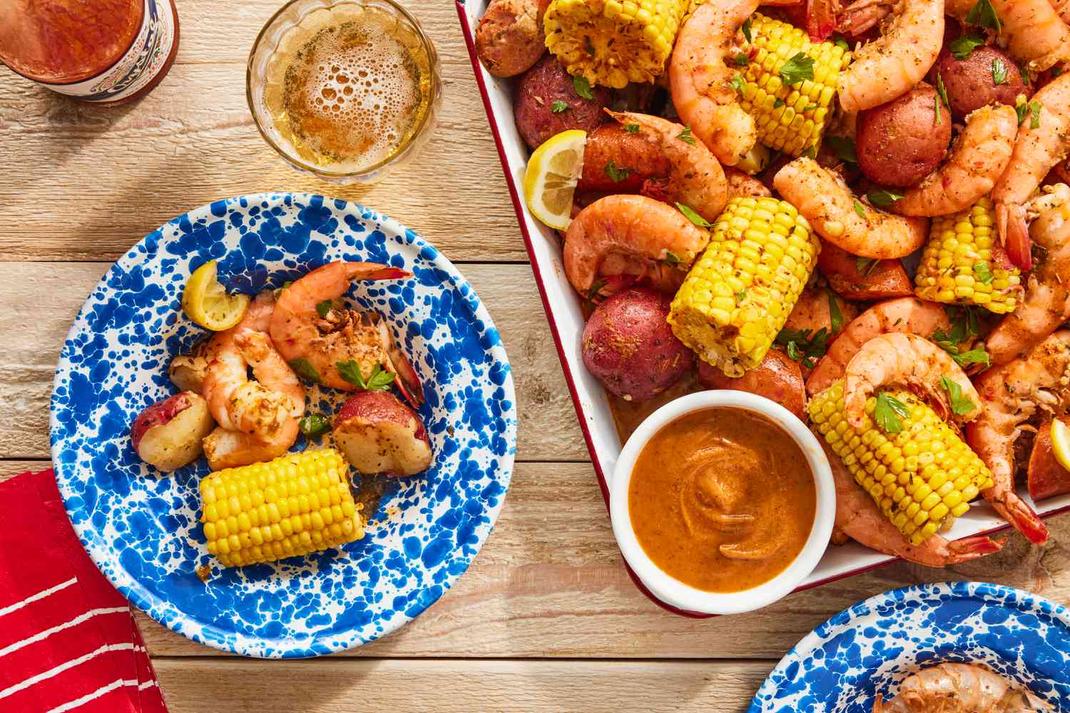 A large platter piled with pink cooked shrimp, halved corn on the cob, baby potatoes, and sliced sausage, scattered with parsley and lemon wedges on a rustic table