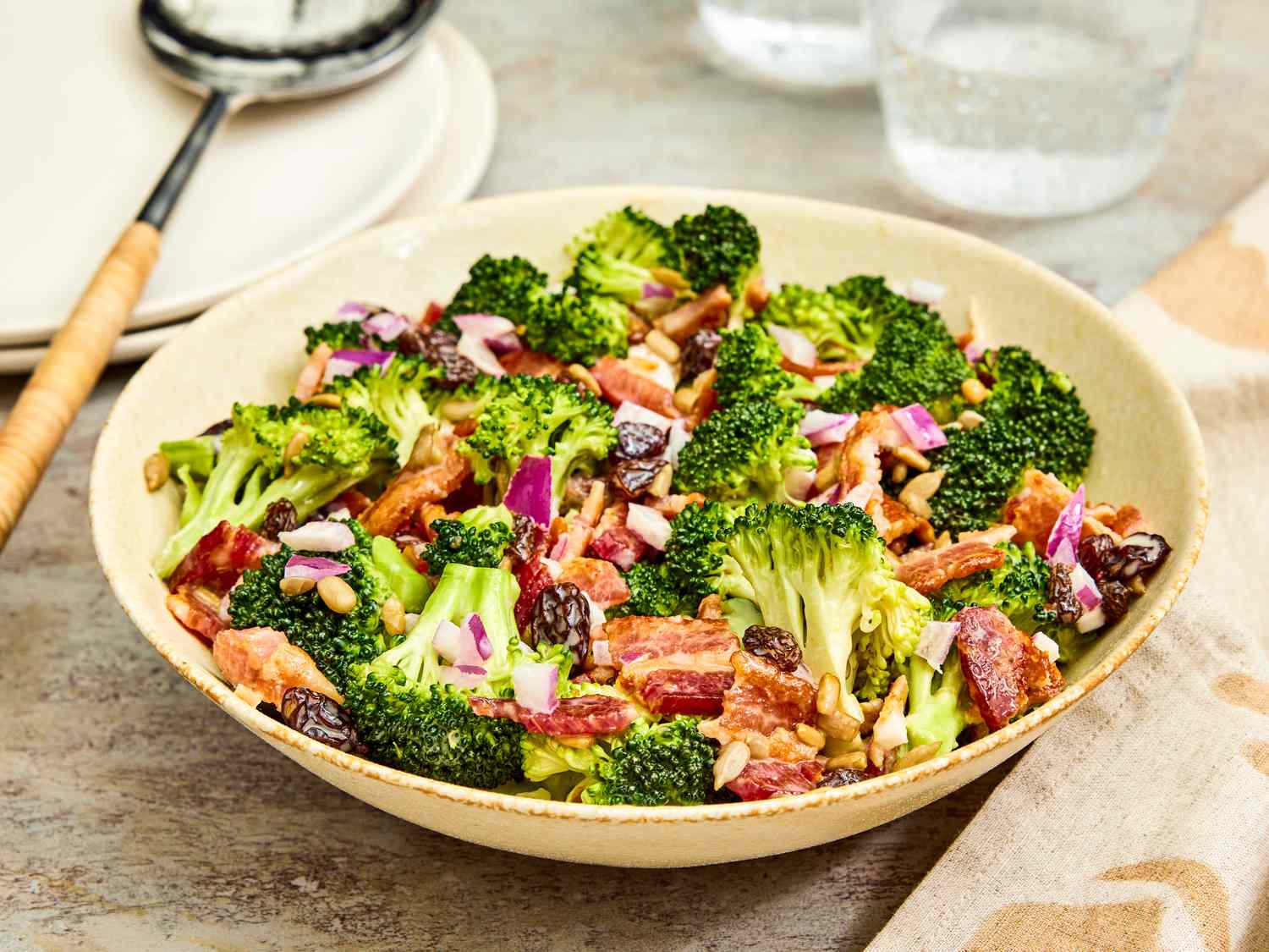 A large serving bowl filled with broccoli salad with bacon, red onion, sunflower seeds, and dried cranberries, creamy dressing lightly coating the ingredients