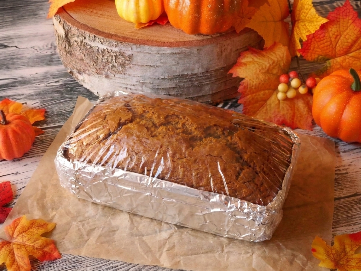 A loaf of pumpkin bread wrapped in plastic wrap and foil on a kitchen counter, ready to freeze