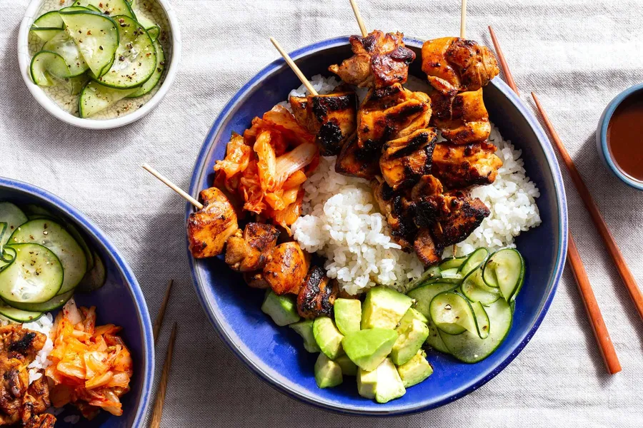 A plated bowl of Korean BBQ chicken thighs sliced and glazed, topped with sesame seeds and sliced scallions, served with steamed rice and cucumber slices on a light wooden table