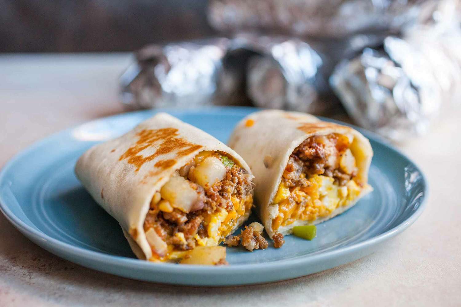 A platter of golden breakfast burritos with one burrito cut open to show eggs, potatoes, and cheese, on a bright kitchen counter