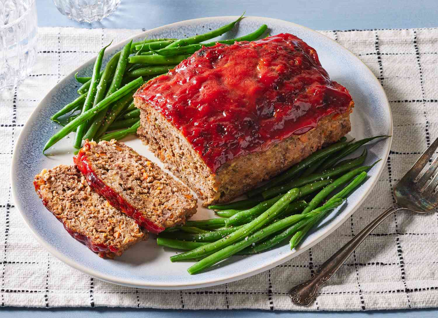 A single loaf of classic meatloaf on a white platter with a shiny ketchup-based glaze, sliced to show a moist interior, set on a cozy dinner table