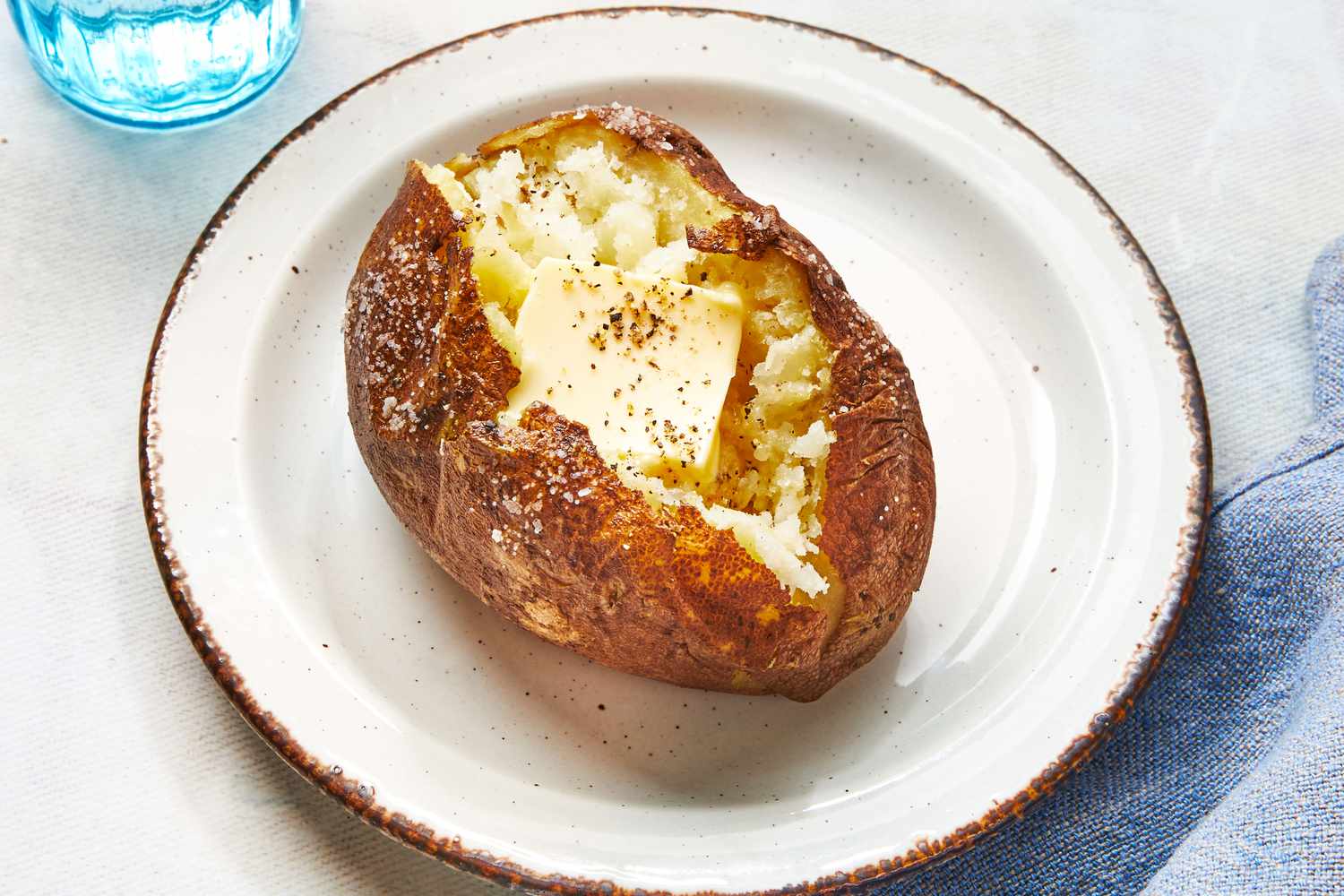 A single oven baked russet potato split open on a plate with a pat of butter melting into the fluffy interior, with crisp salted skin visible