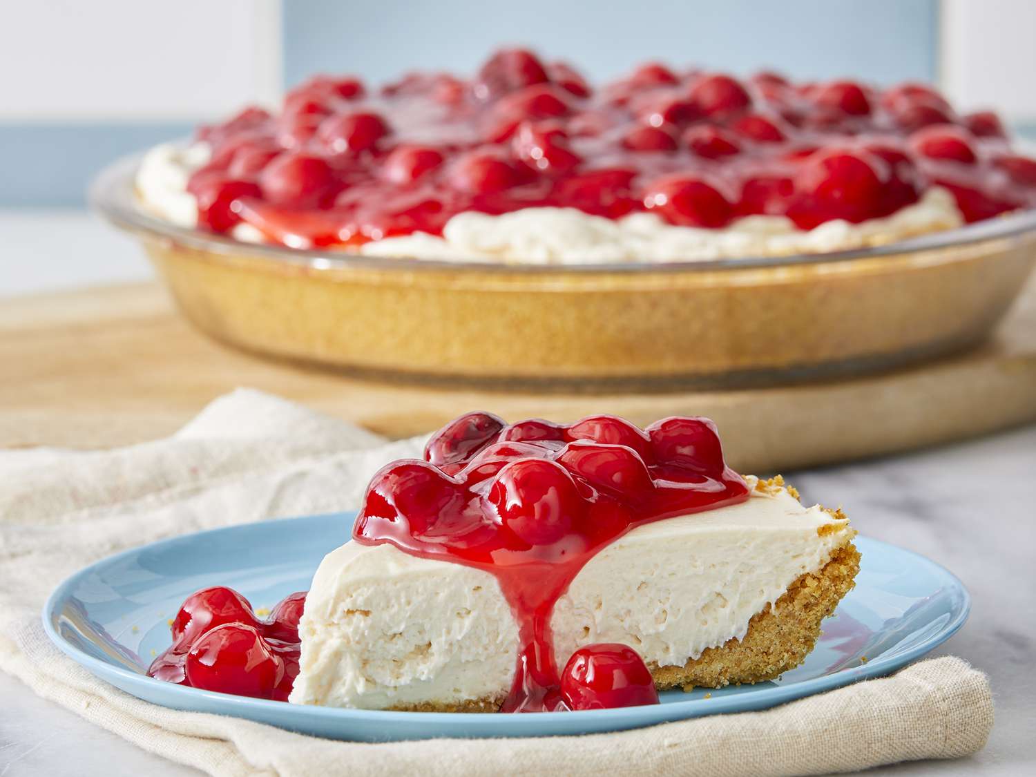 A single slice of no bake cheesecake on a small plate, with a buttery graham cracker crust and a soft, creamy filling, topped with a few fresh berries on a linen-covered table