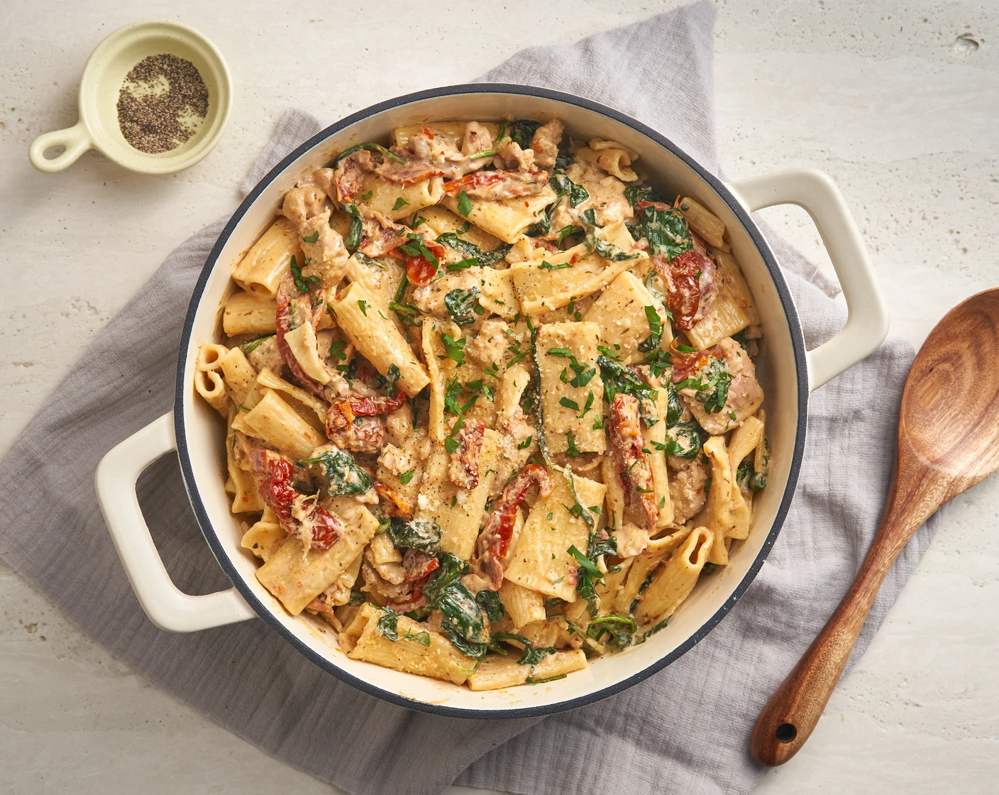 A skillet of creamy Tuscan chicken pasta with sliced chicken, sun-dried tomatoes, spinach, and Parmesan, set on a kitchen counter with a wooden spoon nearby