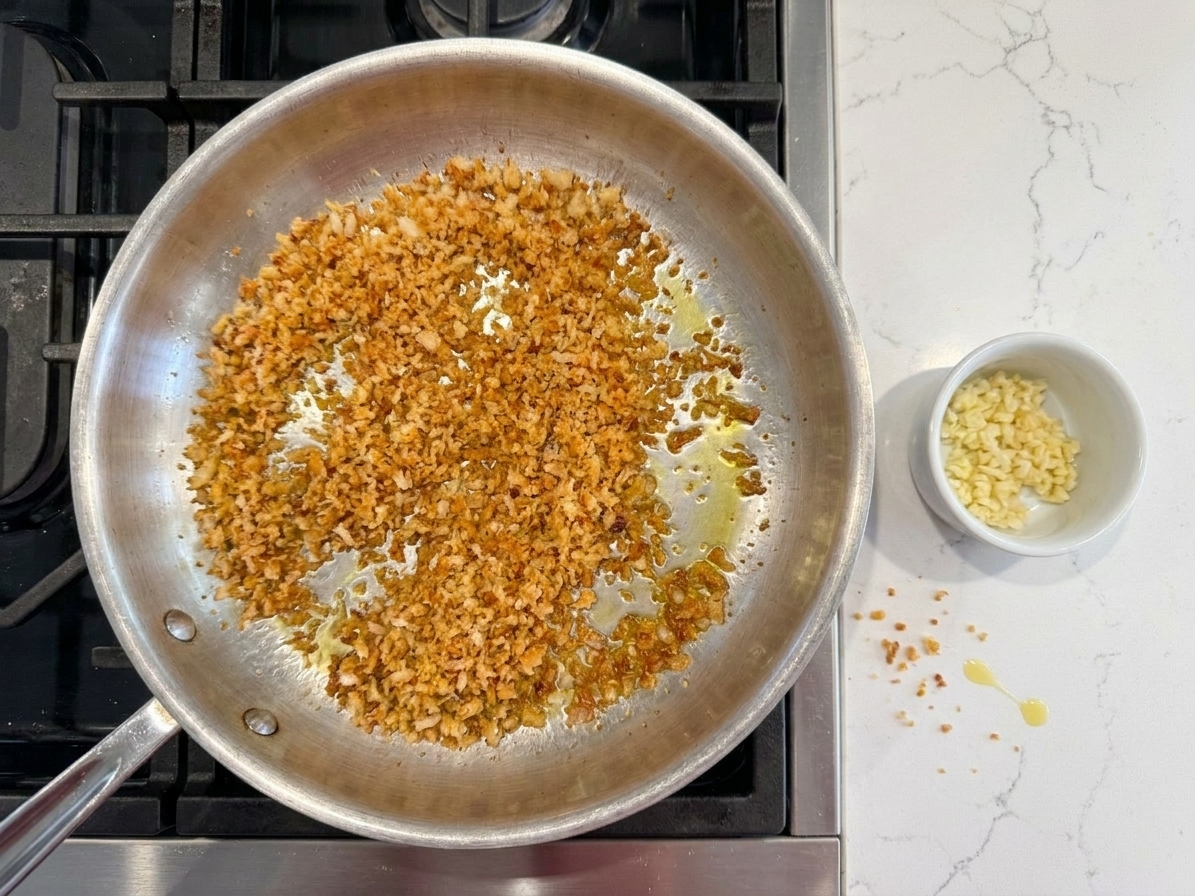A skillet of toasted breadcrumbs turning golden and crisp, with a small pile of minced garlic ready nearby