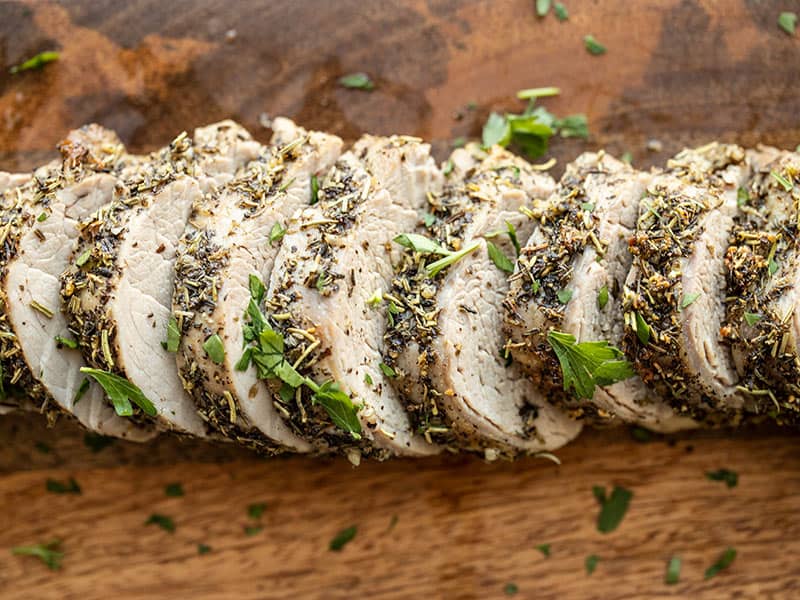 A sliced pork tenderloin on a cutting board with a browned crust and visible juices, garnished with fresh herbs