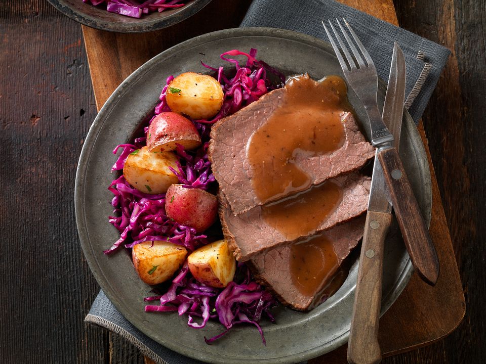 A sliced sauerbraten roast on a platter with rich brown gravy, red cabbage, and potato dumplings on a rustic dinner table
