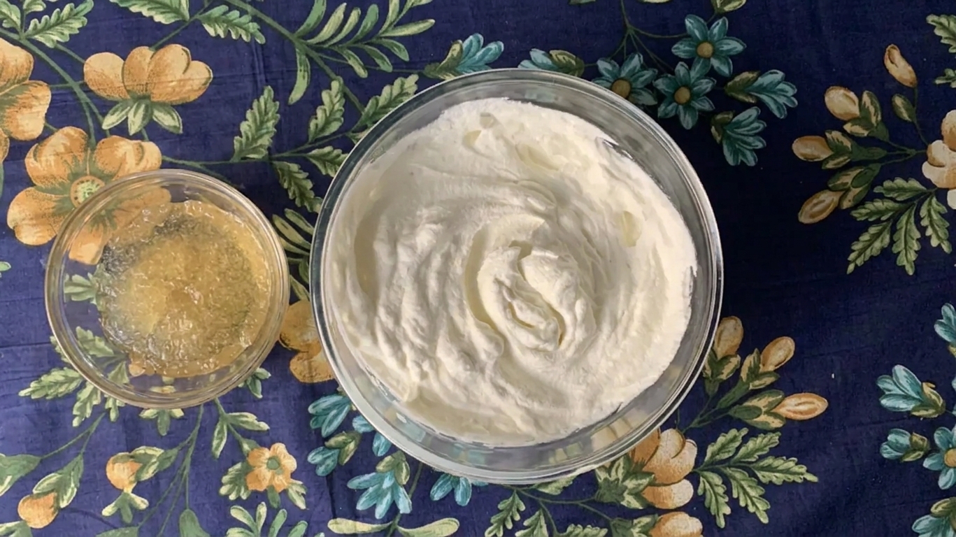 A small bowl of bloomed gelatin beside a mixing bowl of whipped cream on a kitchen counter
