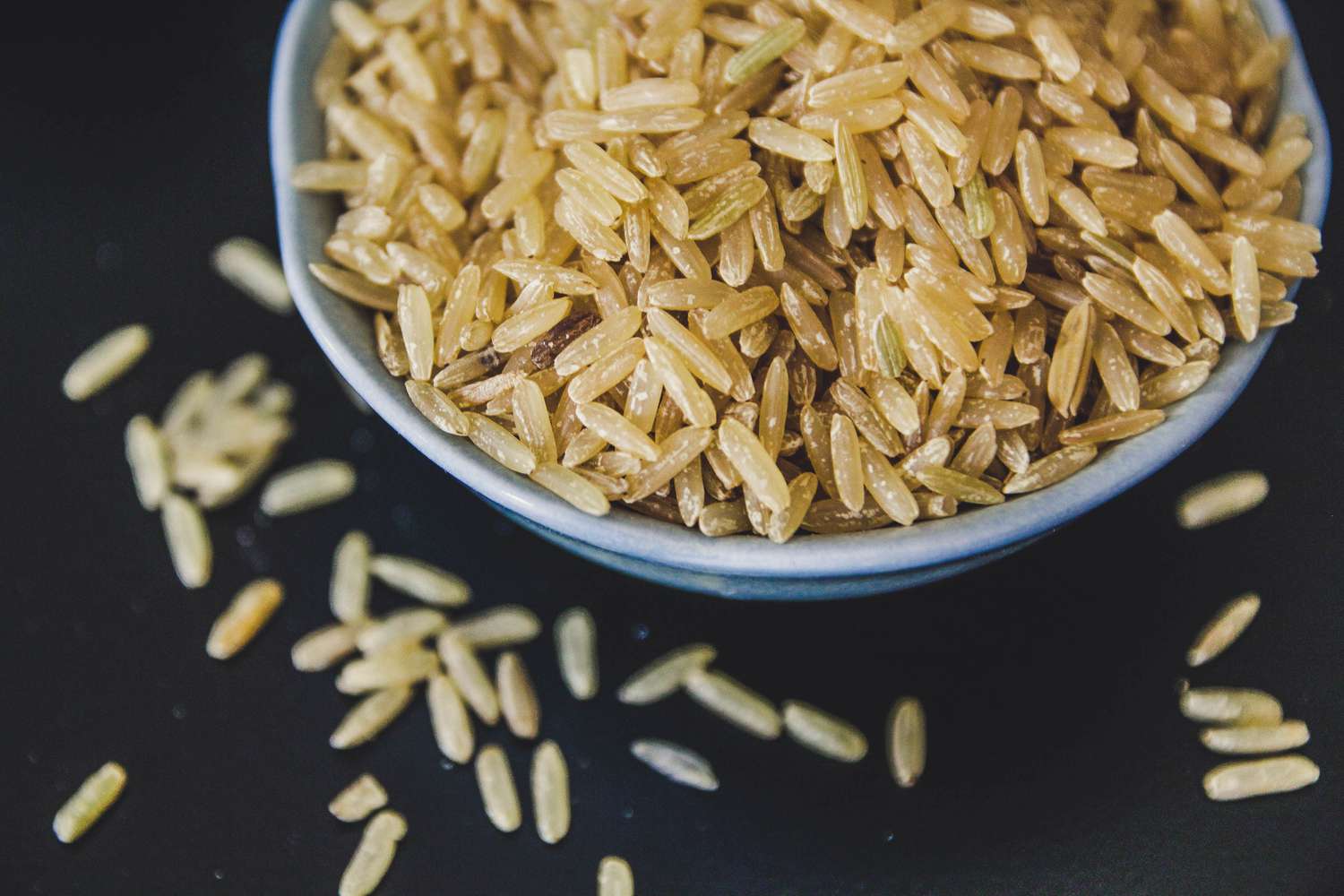 A small ceramic bowl filled with uncooked long-grain brown rice on a wooden kitchen counter with a measuring cup nearby