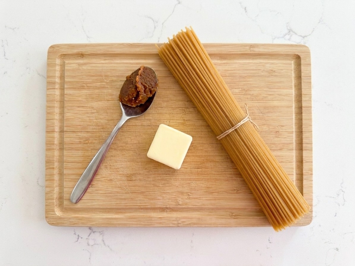 A spoonful of miso paste next to a pat of butter and a bundle of dry spaghetti on a wooden board