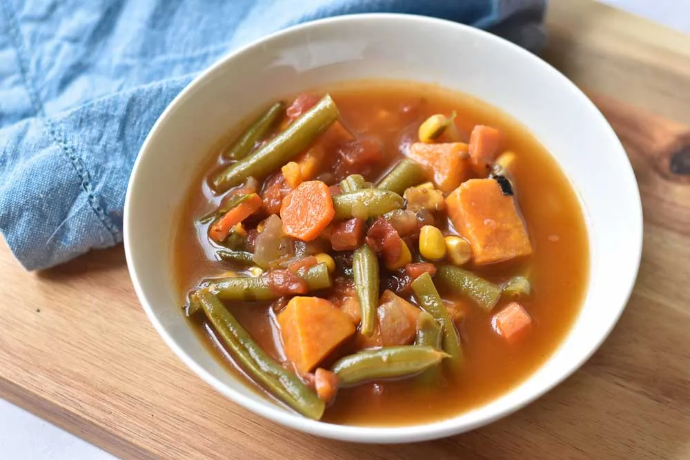 A steaming bowl of garden vegetable soup filled with carrots, zucchini, green beans, tomatoes, and beans in a light tomato broth, sitting on a wooden table with a spoon