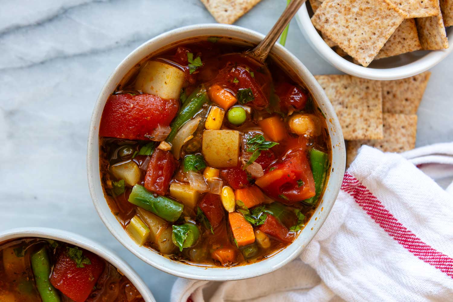 A steaming bowl of hearty vegetable soup with carrots, celery, beans, and greens, served with a piece of crusty bread on a wooden table