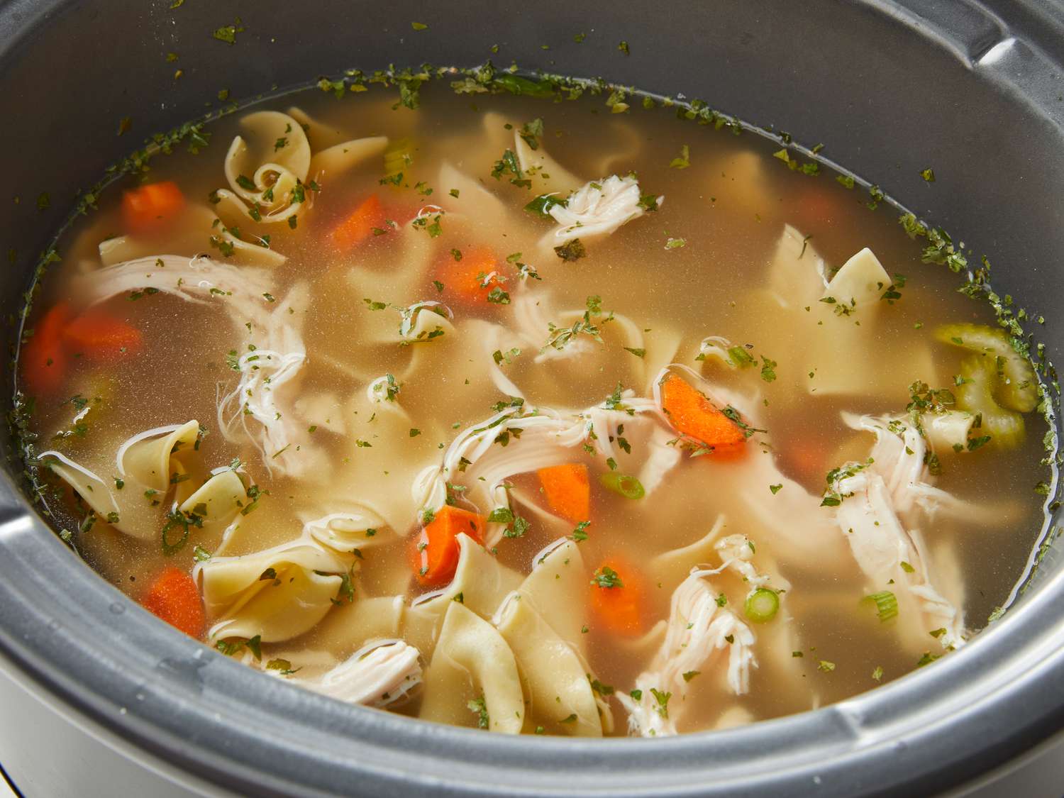 A steaming bowl of slow cooker chicken noodle soup with shredded chicken, sliced carrots, celery, egg noodles, and fresh parsley on top, set on a wooden table with a spoon