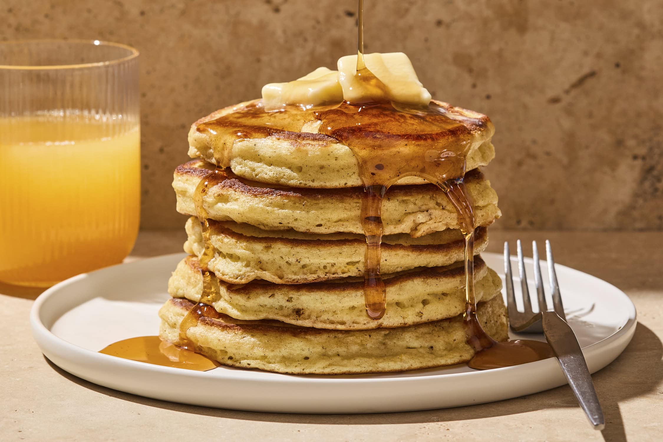 A tall stack of fluffy buttermilk pancakes on a white plate with a pat of butter melting on top and maple syrup dripping down the sides on a sunlit kitchen table