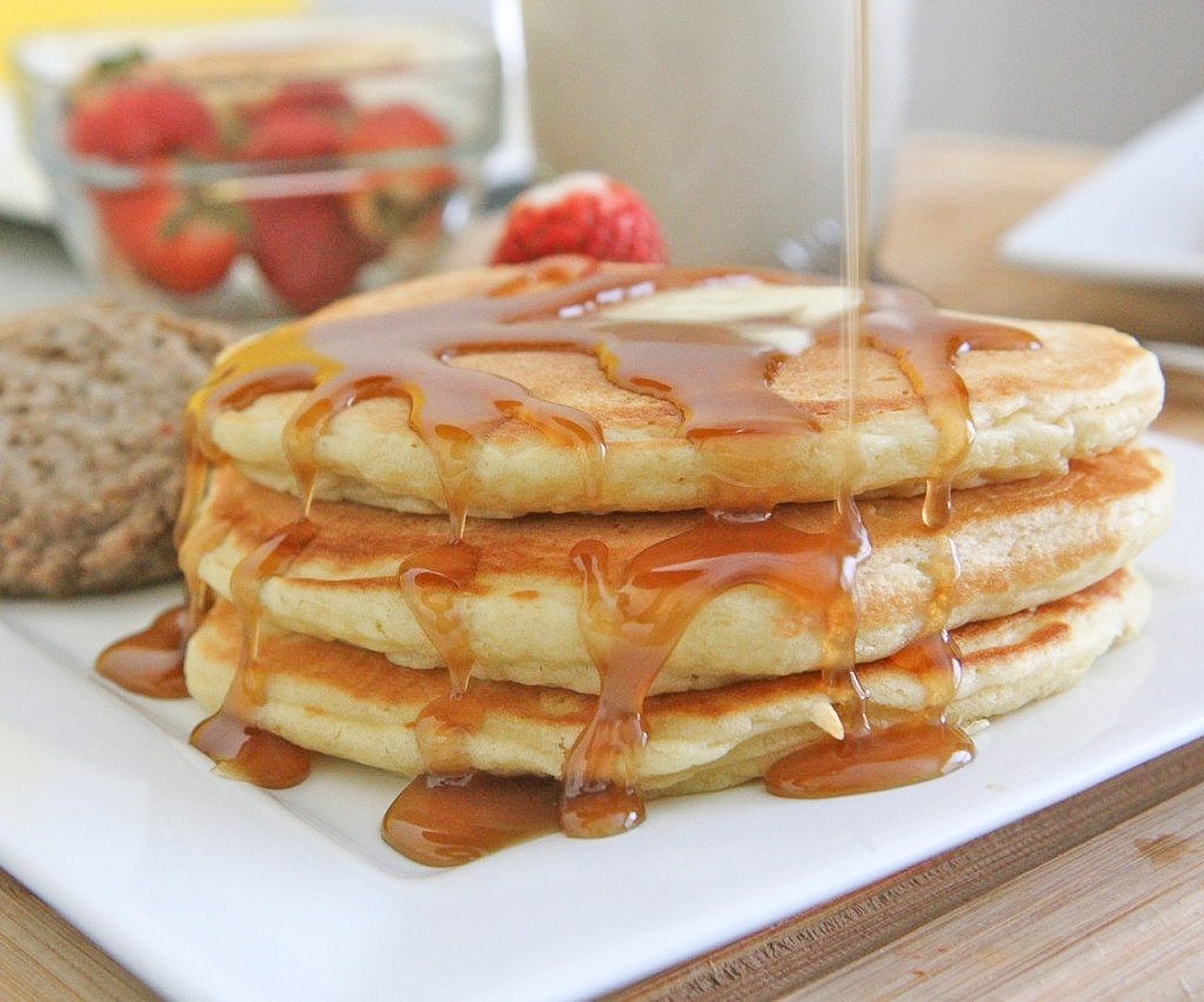 A tall stack of fluffy pancakes on a white plate with a pat of butter melting on top and maple syrup drizzled down the sides on a breakfast table