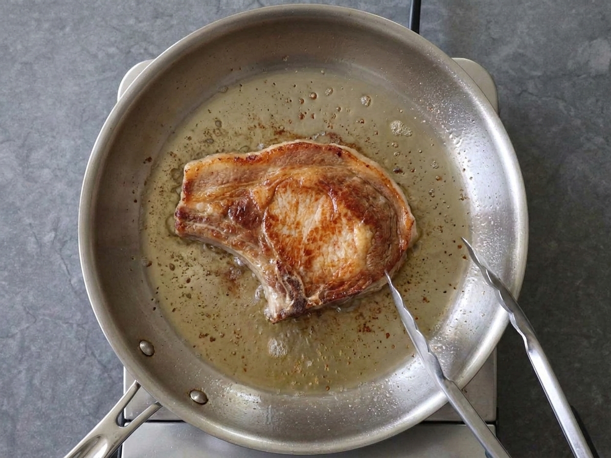 A thick pork chop browning in a stainless steel skillet with sizzling oil and a pair of tongs resting on the pan edge