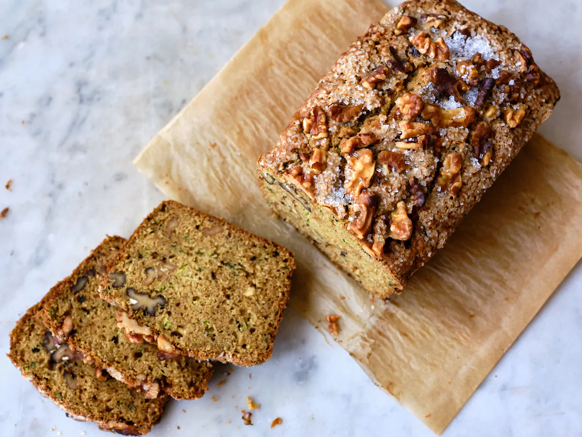 A thick slice of moist zucchini bread on a small plate with visible green zucchini flecks and a tender crumb, set on a wooden kitchen table