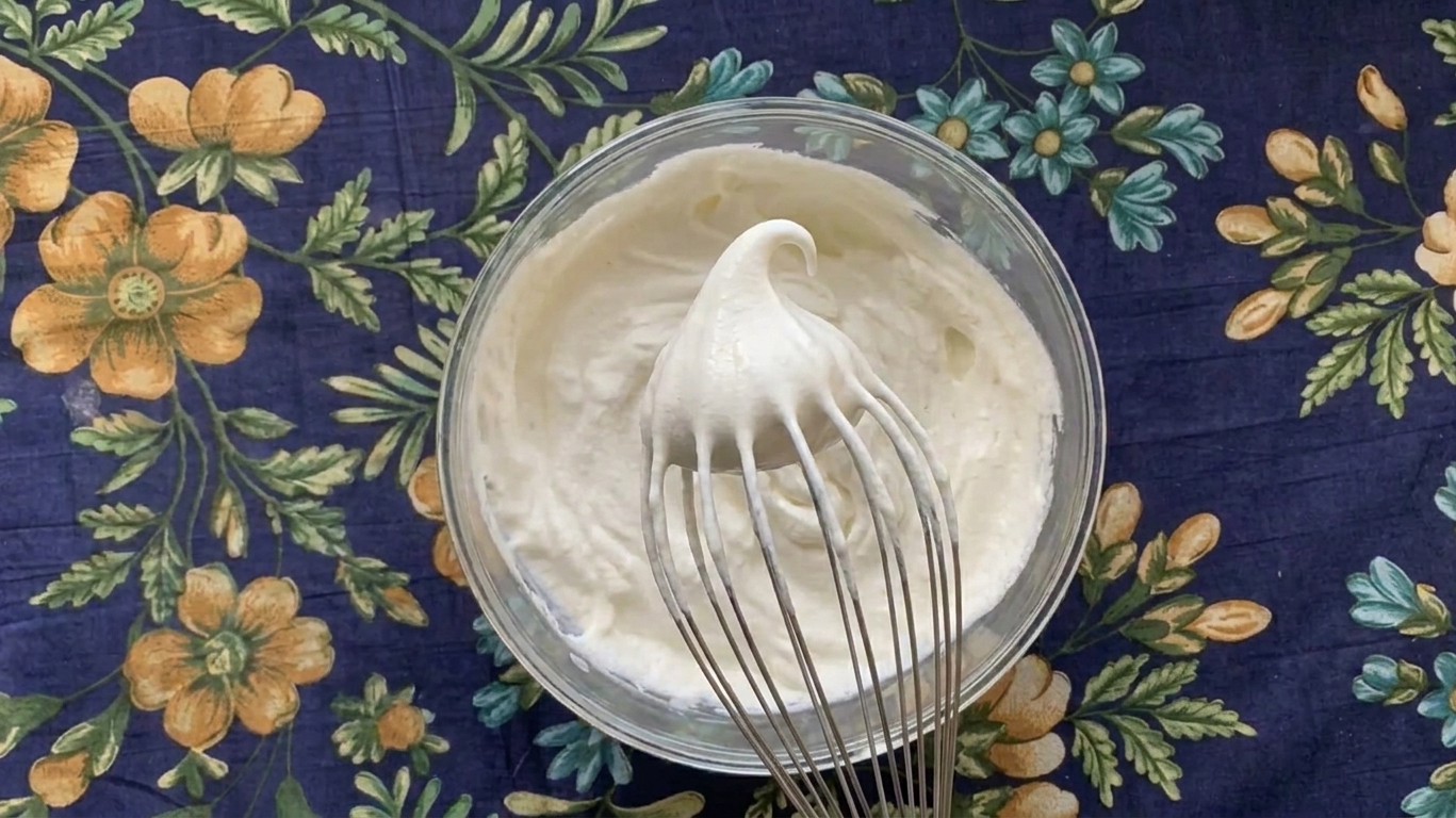 A whisk lifted from a bowl of whipped cream showing a gentle peak that bends over at the tip