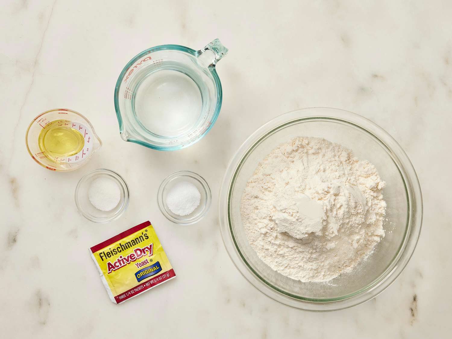 A wooden cutting board on a kitchen counter with flour, instant yeast packets, salt, olive oil, a glass of warm water, and measuring spoons laid out for pizza dough