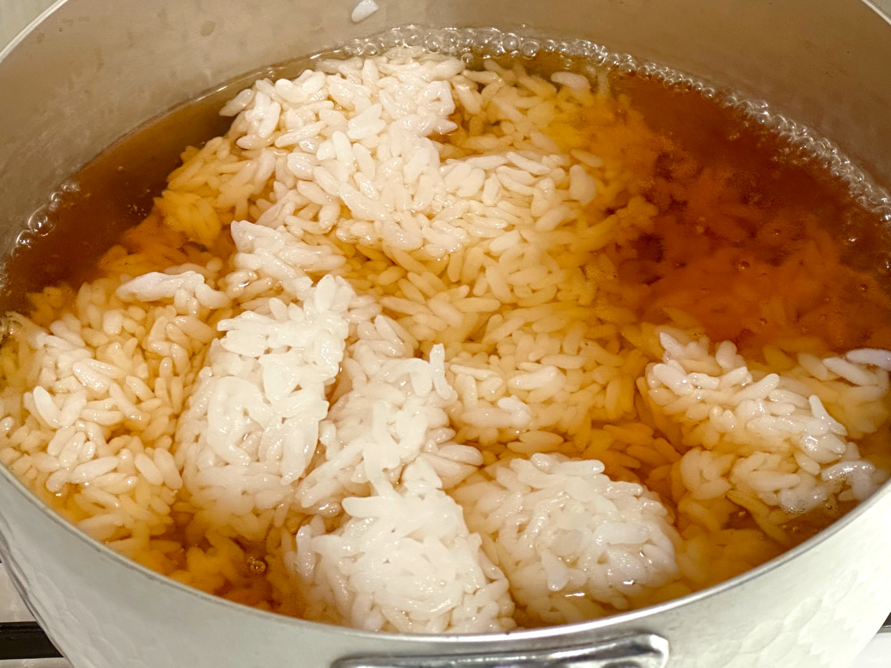 A wooden spoon stirring uncooked rice into broth in a skillet with sautéed onions and garlic, showing a glossy, lemony liquid beginning to simmer