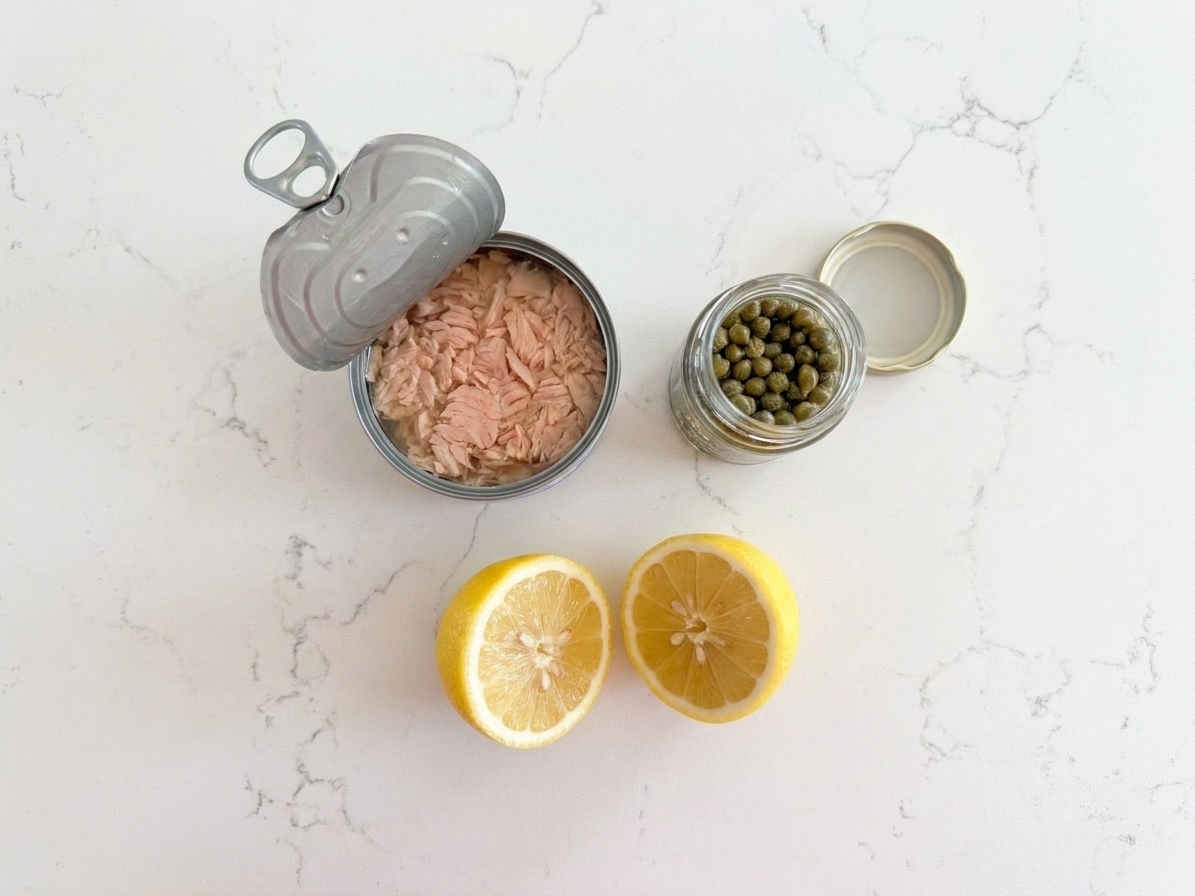 An opened can of tuna, a small jar of capers, and a halved lemon on a kitchen counter