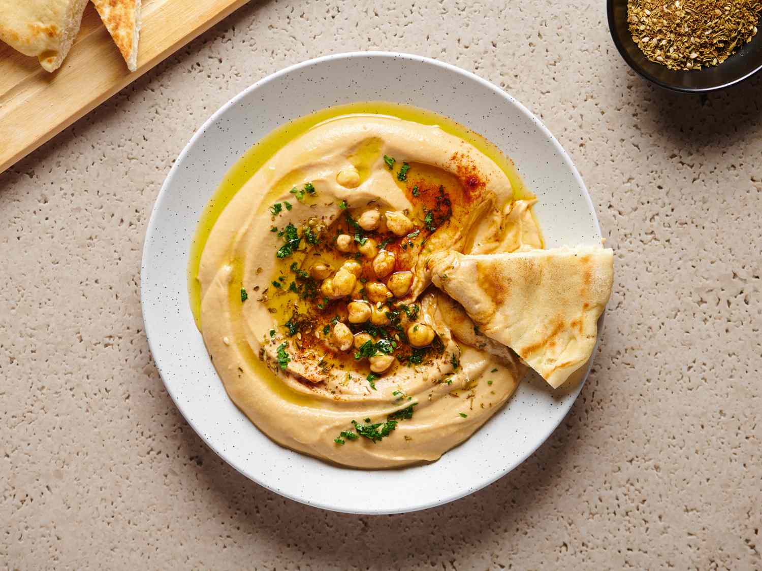 Canned chickpeas, a jar of tahini, fresh lemons, garlic cloves, olive oil, and small bowls of salt and cumin arranged on a light wooden cutting board