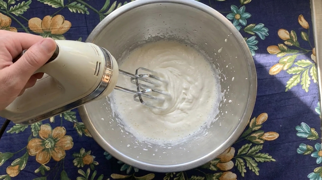 Cream being whipped with a hand mixer in a metal mixing bowl on a kitchen counter