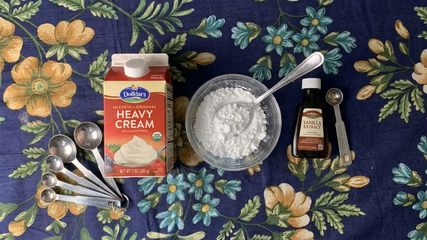 Heavy cream, powdered sugar, and vanilla extract arranged on a kitchen counter with measuring spoons