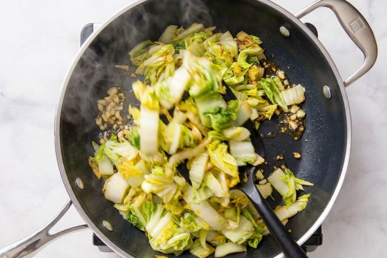 Napa cabbage leaves wilting in a wok as they are tossed with a spatula over a stovetop